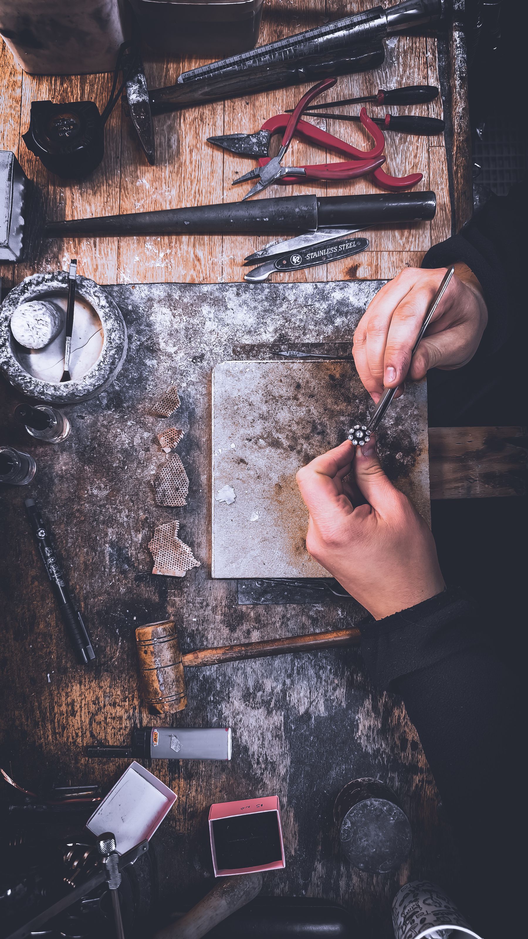 Looking down on a jeweler's bench: hands holding a small piece among pliers, files and hammers.